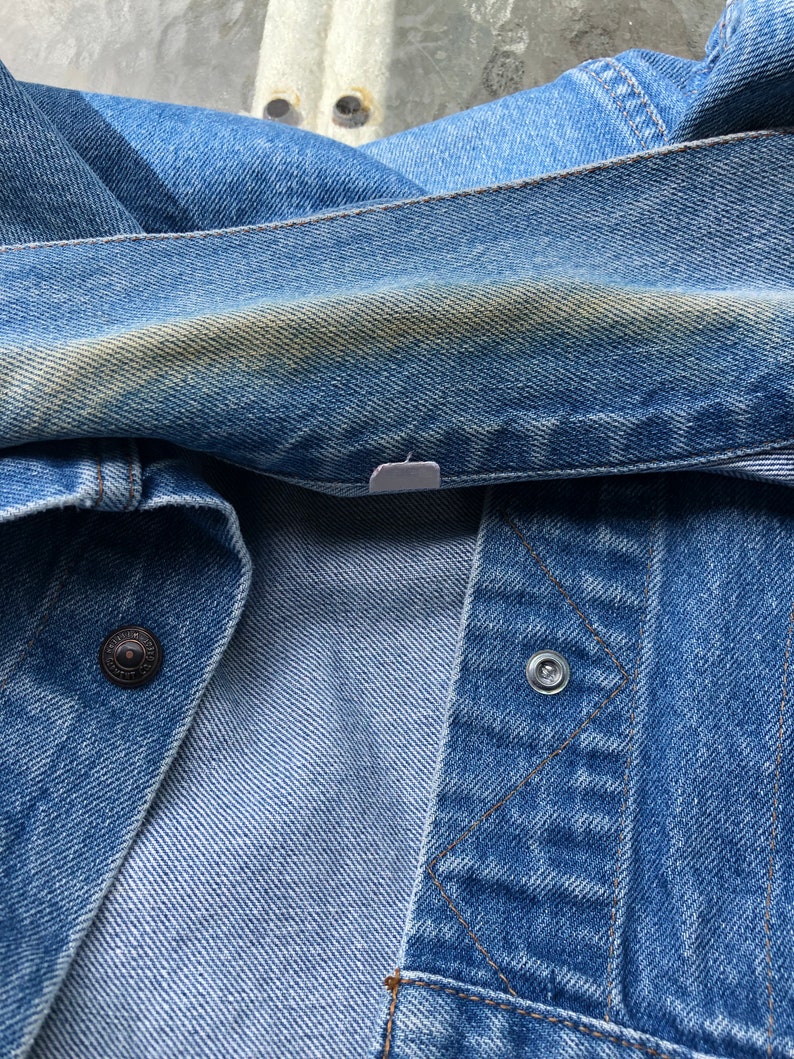 May include: Close-up of a blue denim jacket. The jacket has a button closure and a metal snap. The denim has a faded appearance with some discoloration. The stitching is visible, and the fabric is folded.