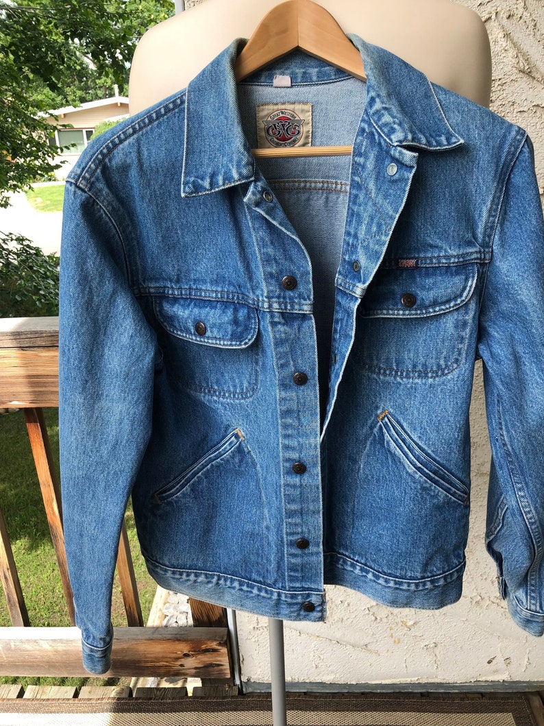 May include: A light blue denim jacket with a classic design, featuring two chest pockets and two side pockets. The jacket has a button-up front and a collar. The brand label is visible on the inside of the collar. The jacket is hanging on a wooden hanger.