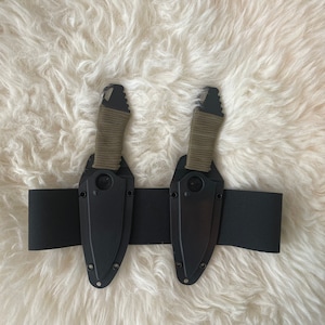 May include: Two black knives with green paracord wraps are attached to a black belt. The knives are on a white fluffy surface.