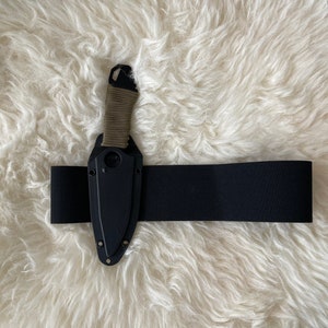 Puede incluir: Un cuchillo negro con mango verde está asegurado en un cinturón elástico negro. El cuchillo y el cinturón están sobre una superficie blanca y esponjosa.