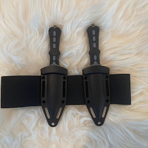 May include: Two black tactical knives with gray accents are sheathed in black leather holsters attached to a black elastic belt.