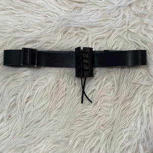 Puede incluir: Cinturón de cuero negro con hebilla metálica y un adorno cilíndrico decorativo con cordones. El cinturón se muestra sobre una superficie blanca y esponjosa.