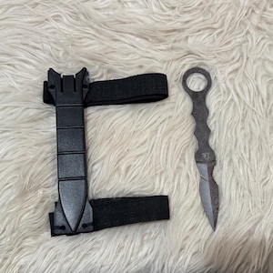 May include: A black arm brace with an elastic strap and a decorative dagger with a patterned blade and handle. The dagger has a ring at the top of the handle.