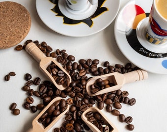 May include: Overhead shot of coffee beans scattered on a white surface, with four small wooden scoops filled with beans. Two espresso cups and saucers are visible, one with a cork coaster. The cups have colorful designs and the saucers have the words "Free time".