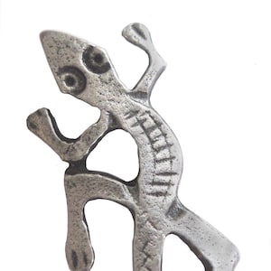 May include: A silver-colored metal lizard pendant with a detailed design. The lizard is in a standing position with its tail curled around its body.