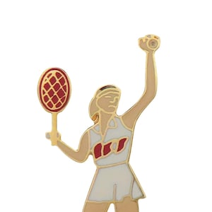 May include: Gold-toned metal pin of a tennis player in a white dress with red accents, holding a tennis racket and a tennis ball.