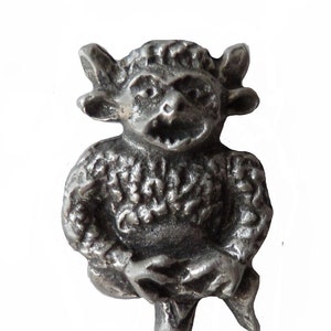 Op de afbeelding: Een zilverkleurig tinnen beeldje van een goblin met een ondeugende uitdrukking. De goblin draagt een getextureerd, gepatroneerd kledingstuk en heeft puntige oren en hoorns.