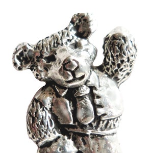 May include: A silver pewter figurine of a teddy bear wearing a shirt and tie. The bear is standing with its right arm raised and its left arm at its side.