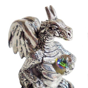 Puede incluir: Una figurita de dragón de plata con las alas extendidas, sosteniendo una gema iridiscente en sus garras. El dragón está sentado sobre una base rocosa.