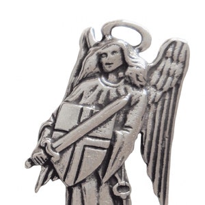 Pode incluir: Uma figura de anjo de prata com asas, segurando uma espada e um escudo. O anjo está vestindo uma túnica e tem uma auréola acima de sua cabeça.