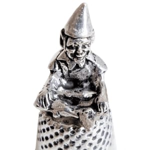 May include: A silver thimble with a detailed gnome figurine sitting on top. The gnome is wearing a pointed hat and has a beard.