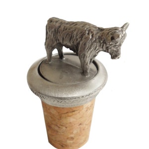 May include: A silver Highland cow bottle stopper with a cork base. The cow is standing on a round base with a textured rim.