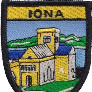 May include: A yellow and black embroidered patch with the word "IONA" in black letters at the top. The patch depicts a yellow building with a blue roof and green grass in front of it.