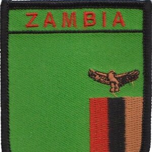 Puede incluir: Un parche bordado verde y negro con la palabra "ZAMBIA" en letras rojas en la parte superior y la bandera de Zambia en la parte inferior. La bandera presenta una franja vertical roja, negra y naranja con un águila negra volando por encima.