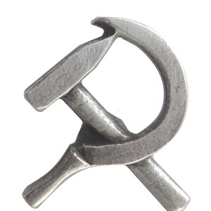 Op de afbeelding: Een zilverkleurige metalen pin in de vorm van een hamer en sikkel.