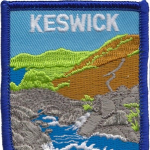 Puede incluir: Un parche bordado azul con la palabra "KESWICK" en letras blancas. El parche presenta una escena de paisaje con colinas verdes, un acantilado marrón y un río azul con olas blancas.