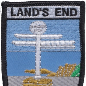 May include: A black and white embroidered patch with the text "LAND'S END" in white. The patch features a white signpost with multiple arms pointing in different directions, a white ball on top, and a yellow base. The signpost is on a blue background with a white sandy beach and green grass.