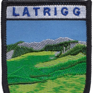May include: Embroidered patch with a blue and white banner that reads "LATRIGG". The patch features a green and yellow landscape with mountains in the background.