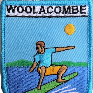 May include: A blue and green embroidered patch with the text "Woolacombe" in white lettering. The patch depicts a cartoon person surfing on a green surfboard in a blue ocean with a yellow sun in the sky.