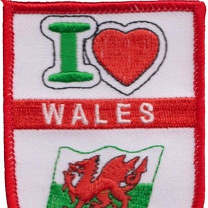 Peut inclure: Un écusson blanc avec une bordure rouge. L'écusson a un cœur rouge avec les lettres "I" en vert et "LOVE" en noir. Sous le cœur se trouve une bannière rouge avec le mot "WALES" en blanc. Le bas de l'écusson présente un fond blanc avec un drapeau vert et blanc du Pays de Galles avec un dragon rouge.
