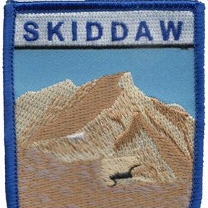 Puede incluir: Un parche bordado azul y blanco con la palabra "SKIDDAW" en letras blancas sobre un fondo azul. El parche presenta una montaña marrón con un cielo azul en el fondo.