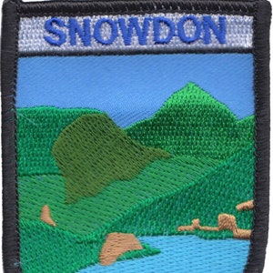 Puede incluir: Parche bordado con fondo azul y la palabra "SNOWDON" en letras azules. El parche representa una cordillera verde con un lago azul en primer plano.