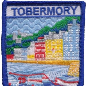 May include: Embroidered patch with a blue border and a white background. The text "TOBERMORY" is in white on the top of the patch. The patch depicts a colorful cityscape with buildings in red, yellow, and blue, and a green hillside in the background. The cityscape is situated on a blue body of water with white boats.