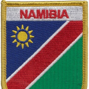 May include: Embroidered patch with the Namibian flag. The flag is a blue field with a gold sun in the upper hoist-side corner. A red diagonal stripe with a white border extends from the lower hoist-side corner to the upper fly-side corner. The lower fly-side corner is green. The patch is edged in gold thread and has the word "NAMIBIA" in red letters at the top.