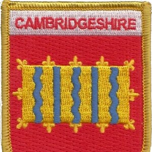 Op de afbeelding: Een geborduurde patch in rood en goud met de tekst "CAMBRIDGESHIRE" in wit. De patch heeft een geel en blauw wapen met een golvend ontwerp.
