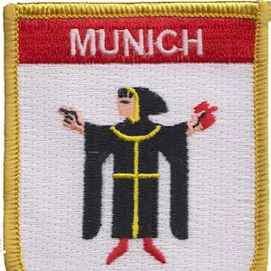 Peut inclure: Un écusson brodé blanc avec une bordure dorée. L'écusson présente le mot "MUNICH" en lettres rouges en haut et une figure noire tenant un objet rouge au centre.