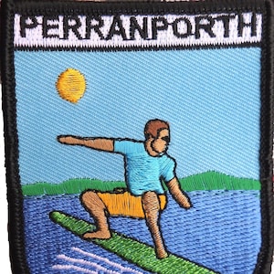 May include: Embroidered patch featuring a surfer riding a wave. The patch has a blue background with a yellow sun and the word "PERRANPORTH" at the top. The surfer is wearing a blue shirt and yellow shorts.