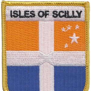 May include: An embroidered patch with a gold border, featuring the text "ISLES OF SCILLY" and a flag with orange, white, and blue stripes and a white star on a blue background.