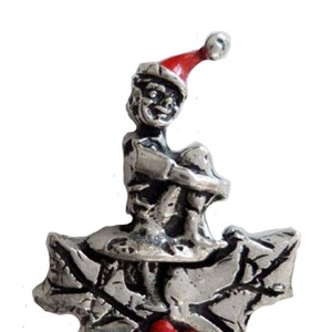 Op de afbeelding: Een zilveren tinnen kerst elf figuur met een rode kerstmanmuts op, zittend op een hulsttak met rode bessen.
