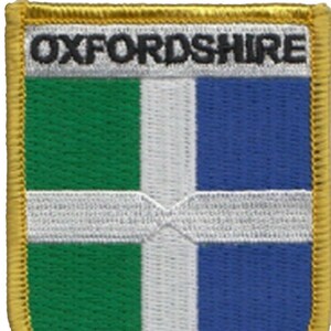 May include: A gold embroidered patch with the text "OXFORDSHIRE" in black lettering. The patch features a white cross on a green and blue background.
