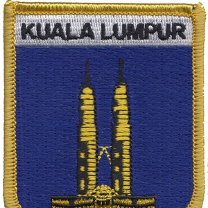 May include: A gold embroidered patch with a blue background featuring the Petronas Towers, a landmark in Kuala Lumpur, Malaysia. The text "KUALA LUMPUR" is embroidered in white on a gray background at the top of the patch.