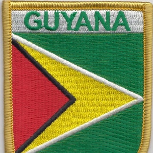 Puede incluir: Un parche bordado con la bandera de Guyana. El parche es verde con una forma de flecha amarilla y roja en el centro. La palabra "GUYANA" está escrita en letras blancas en la parte superior de la bandera. El parche tiene un borde dorado.