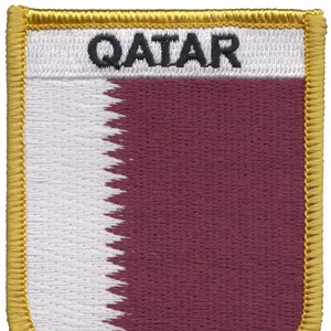 Puede incluir: Un parche bordado en granate y blanco con un borde dorado. El parche presenta la palabra "QATAR" en letras negras.