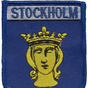 May include: A blue and yellow embroidered patch with the word "STOCKHOLM" in white lettering and a yellow crowned woman's head in the center.