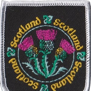 May include: A black embroidered patch with a white border. The patch features three pink thistles with green leaves and stems. The text "Scotland & Scotland" is written in a circle around the thistles.