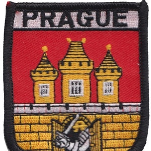 Könnte beinhalten: Ein roter, weißer und gelber gestickter Aufnäher mit dem Wort "PRAGUE" in weißen Buchstaben. Der Aufnäher zeigt ein stilisiertes Bild von drei Türmen mit einem Tor und einer Figur, die ein Schwert hält.