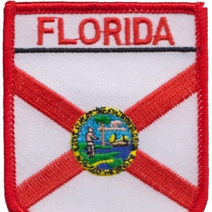 May include: An embroidered patch featuring the state flag of Florida. The patch is red, white and blue with the word "FLORIDA" in red lettering and the state seal of Florida in the centre. The seal depicts a palm tree, a sun and a man standing on a crocodile.