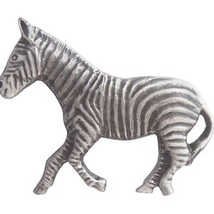 Pode incluir: Uma figura de zebra de estanho prateado. A zebra é representada em uma pose de caminhada com a cabeça virada para o lado. A figura é detalhada com listras e tem um acabamento liso e polido.