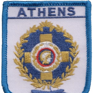 May include: Embroidered patch with a white background and a blue border. The patch features a gold wreath with a blue and gold shield in the center. The shield has a white circle with a red ring and a gold figure inside. The text "ATHENS" is embroidered in blue at the top of the patch.