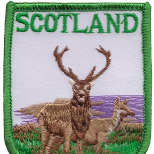 Puede incluir: Parche bordado con borde verde y fondo blanco. El parche presenta la palabra "SCOTLAND" en letras verdes en la parte superior. El centro del parche representa un ciervo marrón con grandes cuernos de pie frente a un paisaje morado y blanco.