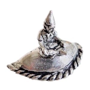 Könnte beinhalten: Eine silberne Figur eines Zwergs, der auf einem Gebäck sitzt. Der Zwerg trägt einen spitzen Hut und hat ein detailliertes Gesicht. Das Gebäck ist halbmondförmig und hat eine geflochtene Kruste.