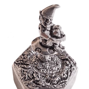 Puede incluir: Una caja de alhajas de peltre plateado con un diseño detallado de un gnomo sosteniendo una pala. La caja es hexagonal y tiene una tapa con bisagras.