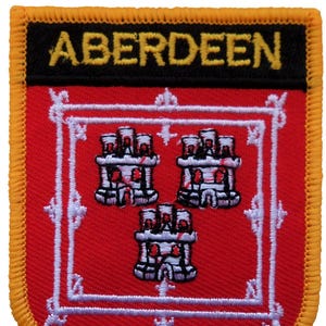 May include: Embroidered patch featuring the word "ABERDEEN" in yellow letters on a black banner. The patch has a red background with three white and red castle emblems. A white decorative border and yellow stitching complete the design.