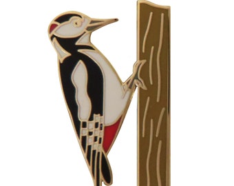 May include: A gold-colored enamel pin featuring a black and white woodpecker perched on a brown tree trunk. The woodpecker has a red underbelly and a black and white checkered pattern on its tail feathers.