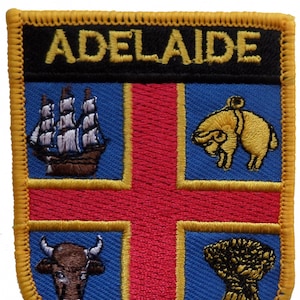Op de afbeelding: Geborduurde patch met het woord "ADELAIDE" bovenaan. Het ontwerp omvat een schip, ram, stier en tarwe. De achtergrond is blauw met een rood kruis en een gele rand.