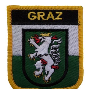 Op de afbeelding: Geborduurde patch met het woord "GRAZ" in goud boven een schild. Het schild toont een witte leeuw met een gouden kroon en rode details op een groene achtergrond. De patch heeft een zwart, wit en groen ontwerp met een gele rand.